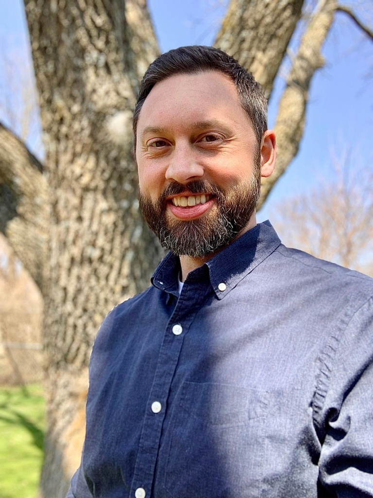 man with beard in button-down shirt standing in front of a tree