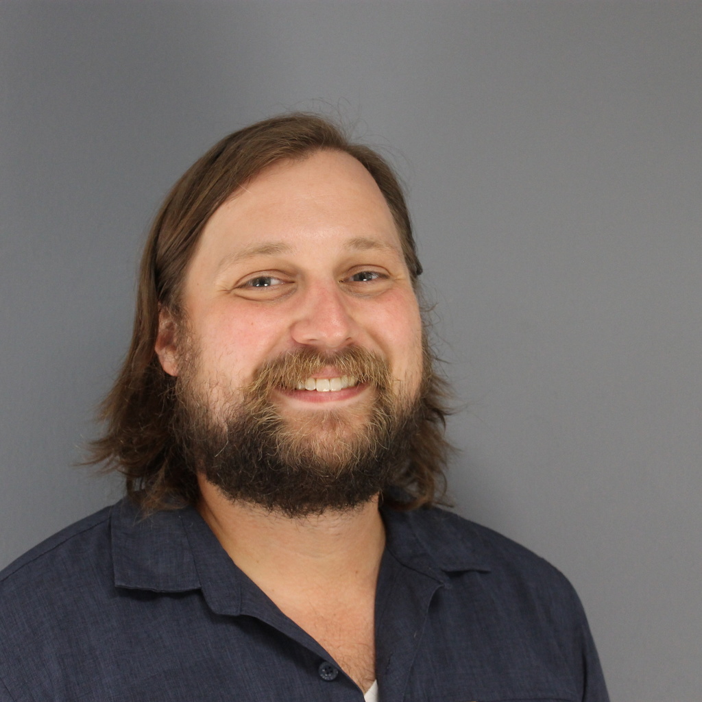 man with beard and longish brown hair wearing a black shirt standing in front of a gray wall